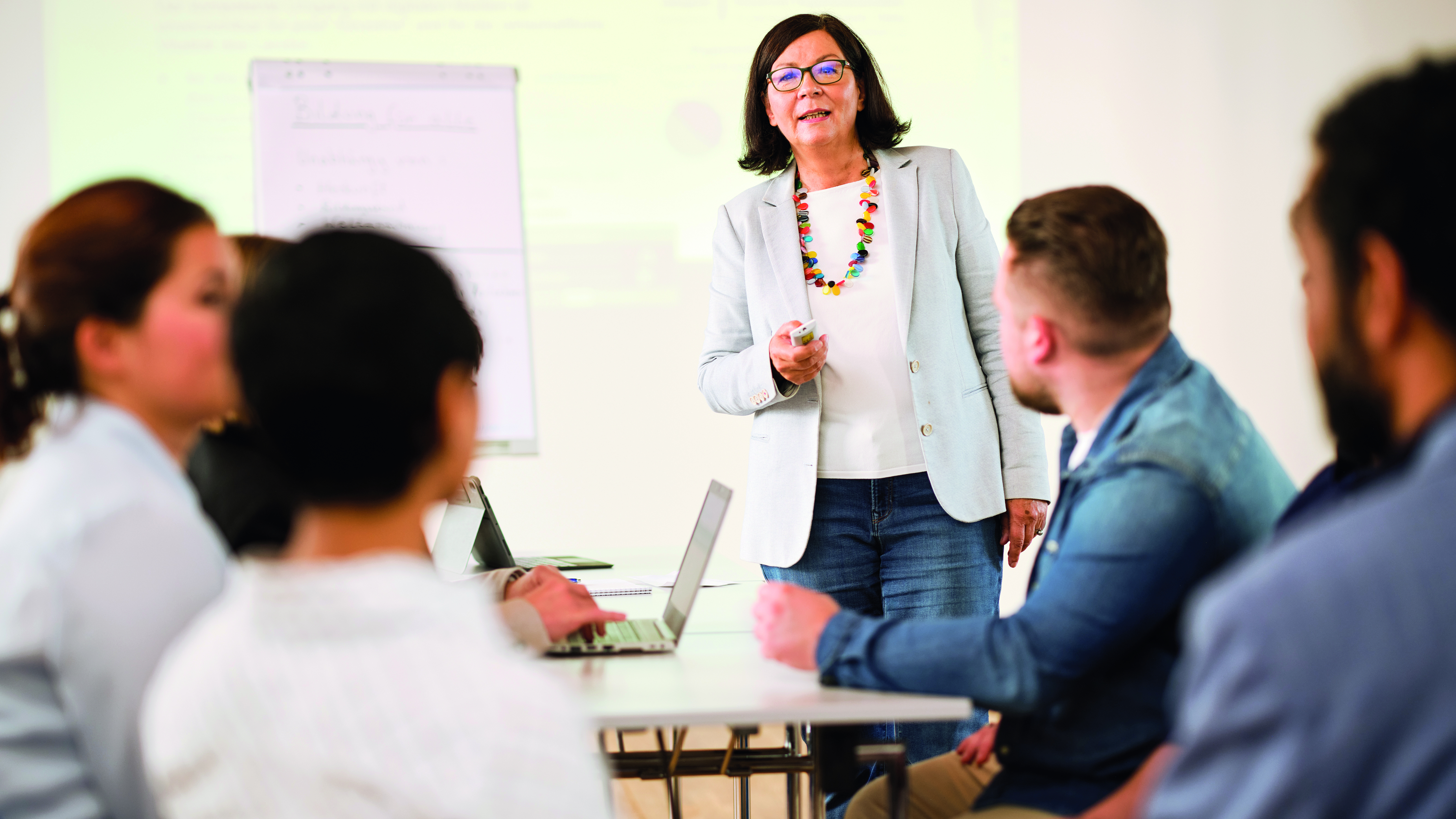 Unterrichtssituation mit unscharfem Vordergrund und fokussierter Lehrperson vor Flipchart.