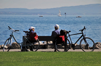 Mann und Frau sitzen auf einer Bank und blicken auf einen See. Es sind Fahrräder zu sehen.