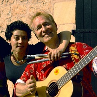 Josephine Kronfli und Pit Budde vom Karibuni-Mitmachkonzert stehen im Sonnenschein vor einer Wand und lächeln in die Kamera. Sie halten Musikinstrumente in den Händen: eine Gitarre und verschiedene Flöten.