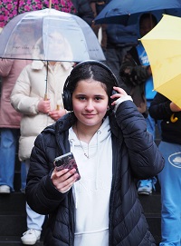 Ein Mädchen mit Kopfhörern und Smartphone blickt lächelnd in die Kamera. Hinter gehen Menschen mit Regenschirmen.