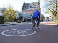 Fahrradstraße mit rotem Asphalt und rechts im Bild ein Fahrradfahrer mit einer blauen Jacke bekleidet.