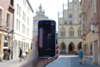 Auf einem Handy ist eine Thermometer-App geöffnet, die 31 Grad Celsius anzeigt. Im Hintergrund ist der Prinzipalmarkt in Münster zu sehen.