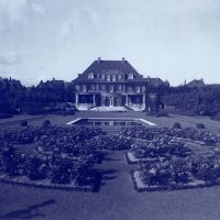 Auf dem Foto sieht man die Villa ten Hompel.
Links und rechts vom Haus stehen Bäume.
Davor ist ein Teich und viele Rosen.
Das Haus und der Garten sind sehr groß.