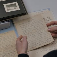 Eine Person sitzt an einem Schreibtisch und hält einen alten Feldpostbrief in der Hand, andere liegen auf dem Tisch neben einem aufgeschlagenen Fotoalbum.