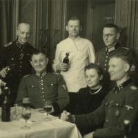 Auf dem Schwarz-Weiß-Foto sitzen und stehen Personen an einem gedeckten Tisch, auf dem Biergläser stehen. Einige haben Bierflaschen in der Hand. Vier Personen tragen Uniform.