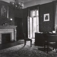 Historische Ansicht eines Arbeitszimmers mit Teppich auf dem Boden, auf dem ein breiter dunkler Schreibtisch mit zwei Stühlen davorsteht. Über einem Kamin hängt ein Gemälde Hitlers.