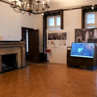 Im Ausstellungsraum befinden sich Tafeln und ein Touchscreen auf einem gekippten Schreibtisch. An einer Raumseite befindet sich der historische Kamin, in der Mitte hängt ein Kronleuchter.