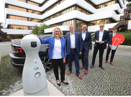 Fünf Personen stehen auf dem Parkplatz vor einem Gebäude neben einer E-Ladesäule