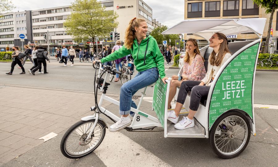 Eine Frau fährt eine Leeze, in der hinten zwei weitere Frauen sitzen, die durch die Stadt gefahren werden