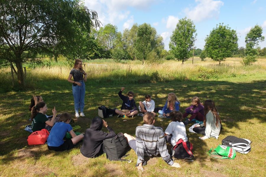 Eine Gruppe Menschen sitzt in einem Feld im Kreis, eine von ihnen steht und die anderen schauen zu ihr