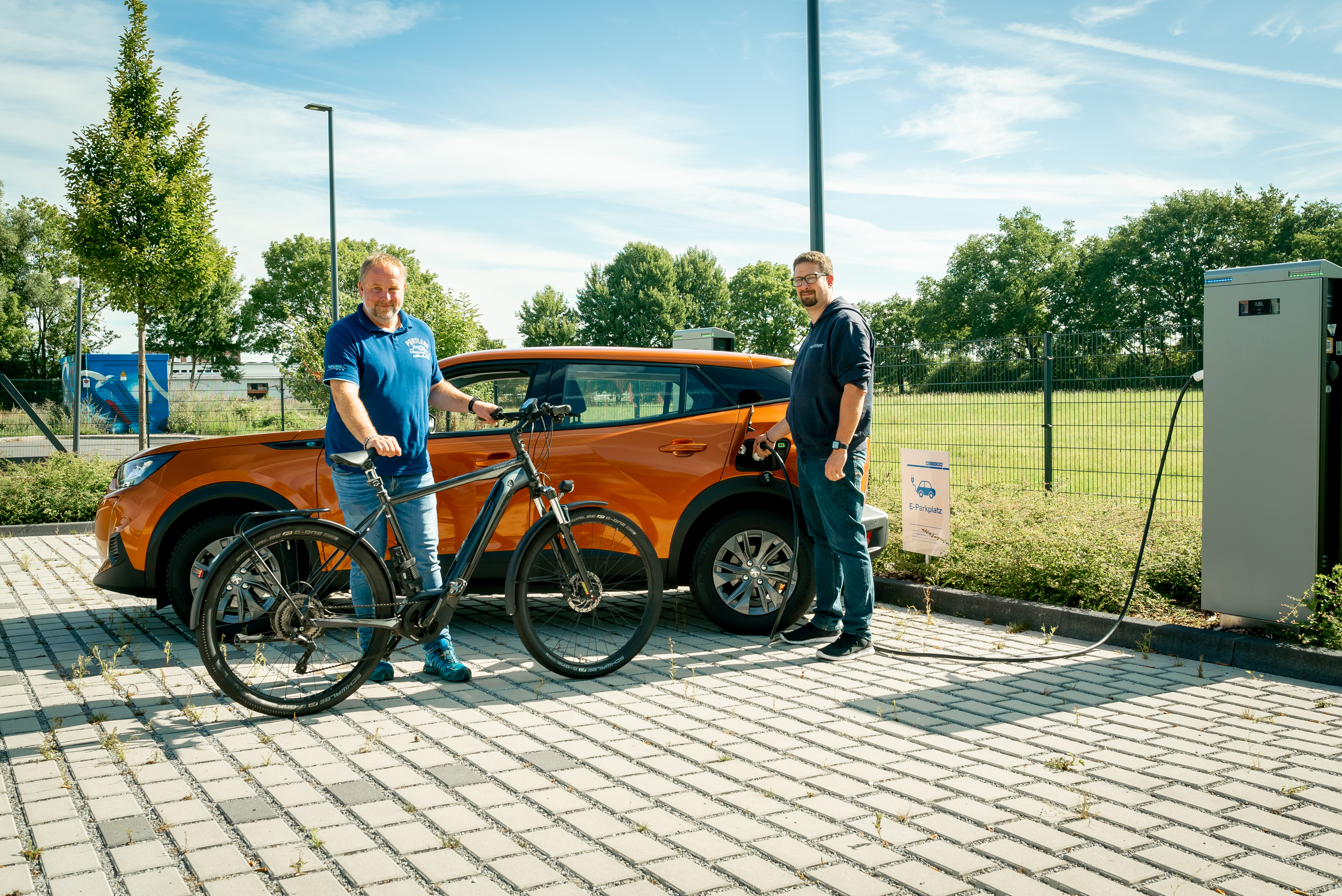 Zwei Männer stehen auf einem Parkplatz, einer kommt mit dem Fahrrad, der andere steht neben seinem Auto, das an einer E-Ladesäule hängt